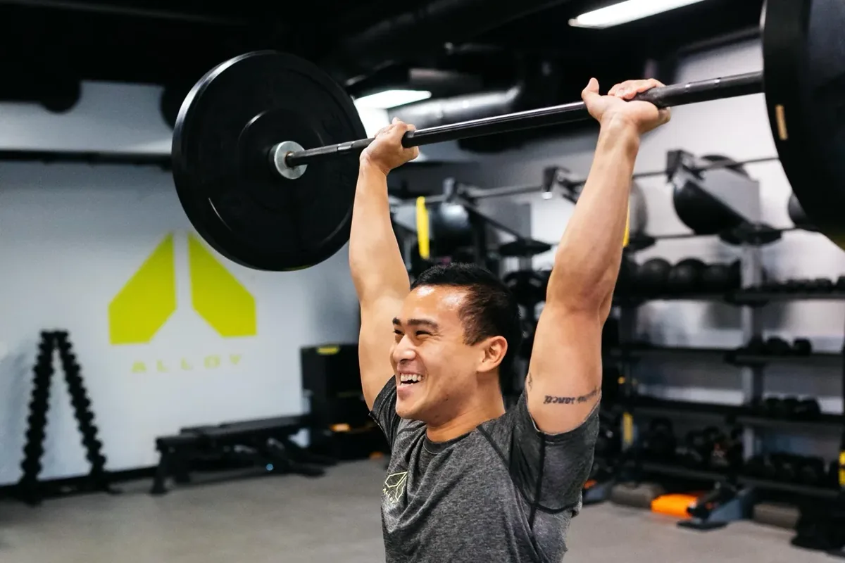 A member presses a loaded barbell overhead during a coached session at an Alloy Personal Training studio