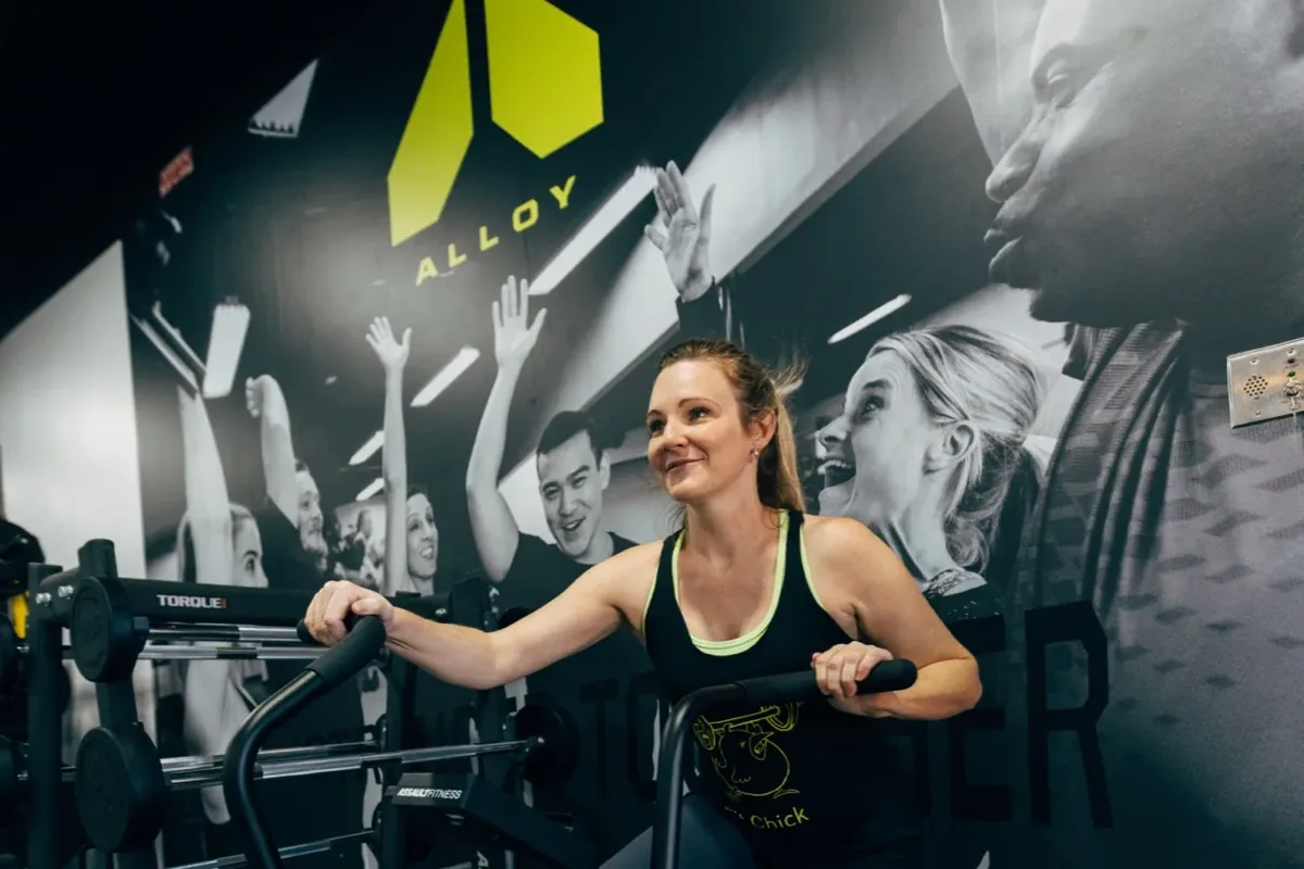 A smiling woman pushes a weighted training sled during a coached session at an Alloy Personal Training studio
