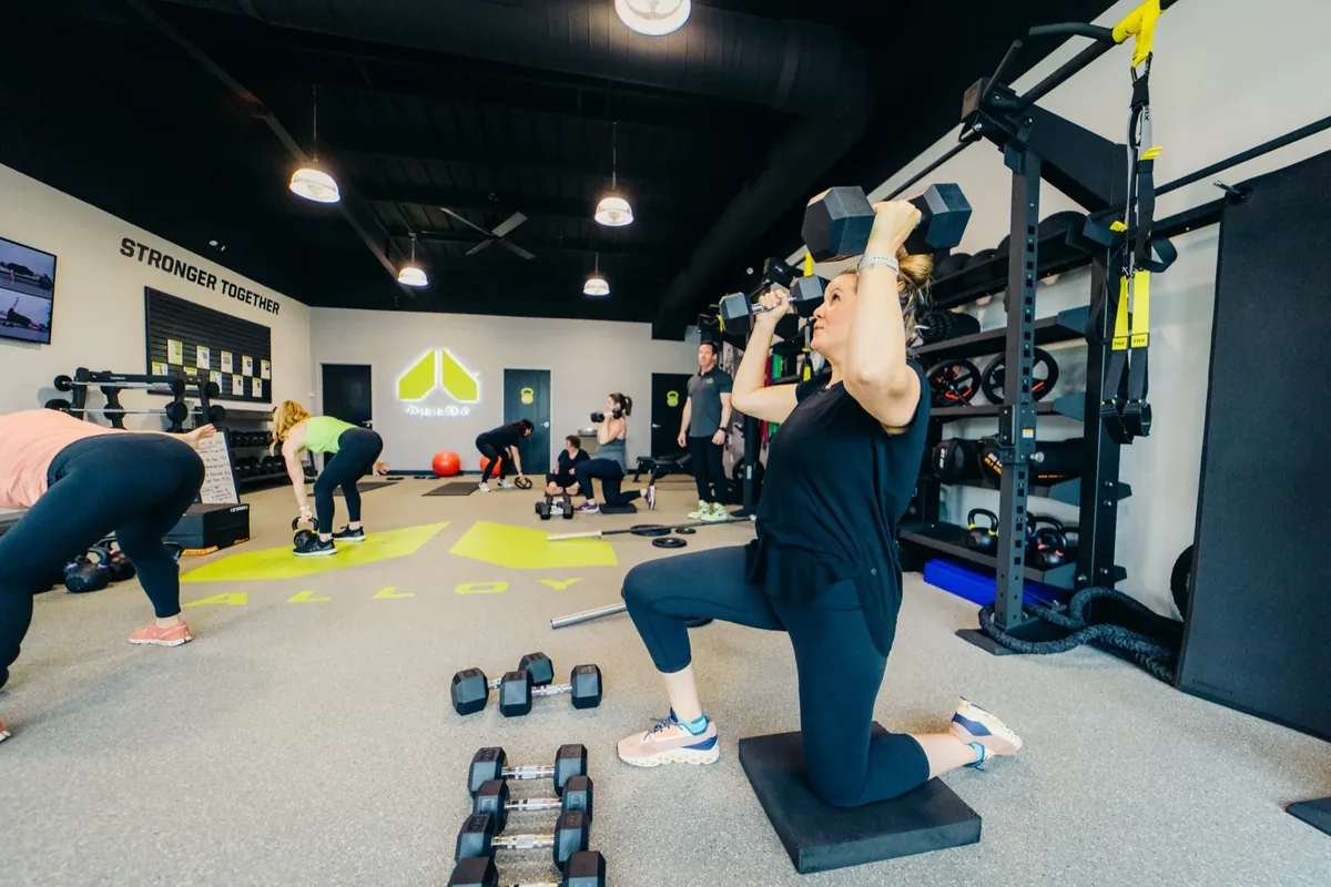 A member presses a dumbbell overhead from a kneeling position while others follow their own programs behind her at Alloy Personal Training Middleton in Middleton, WI.