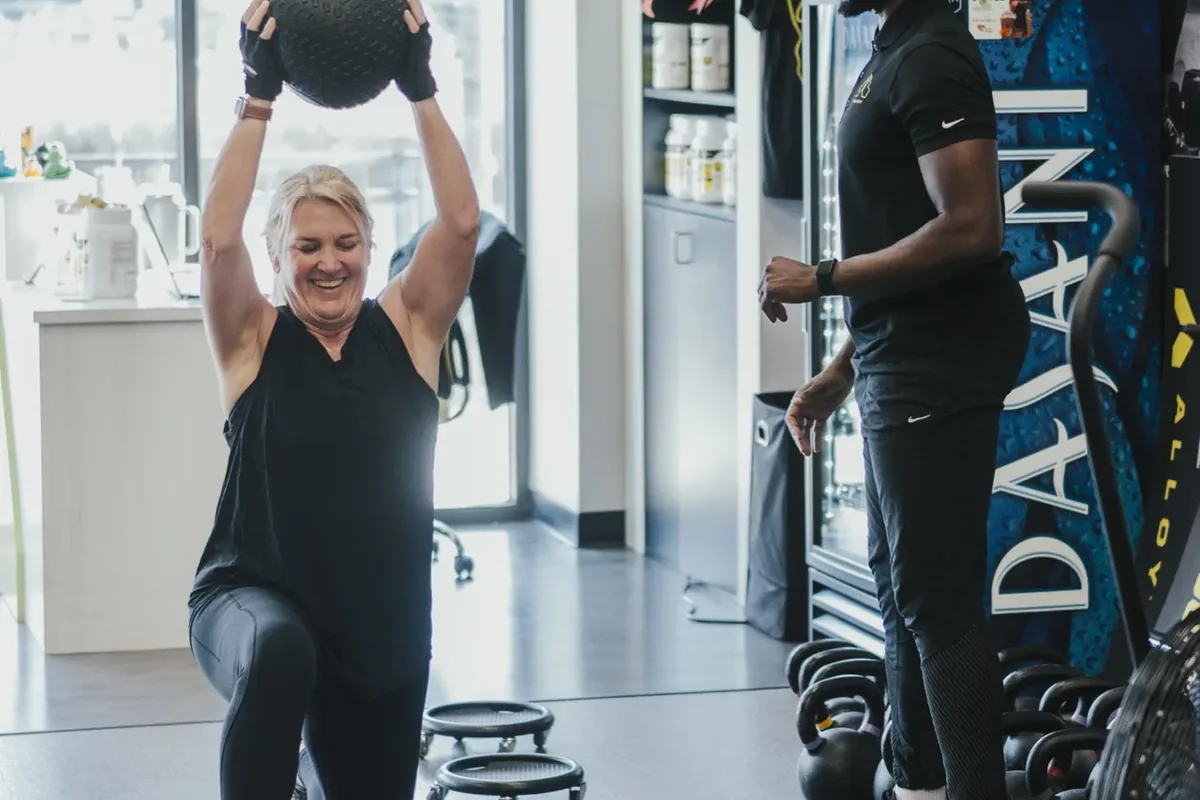 A coach watches a member lunge while pressing a slam ball overhead during a small group session at Alloy Personal Training Middleton in Middleton, WI.