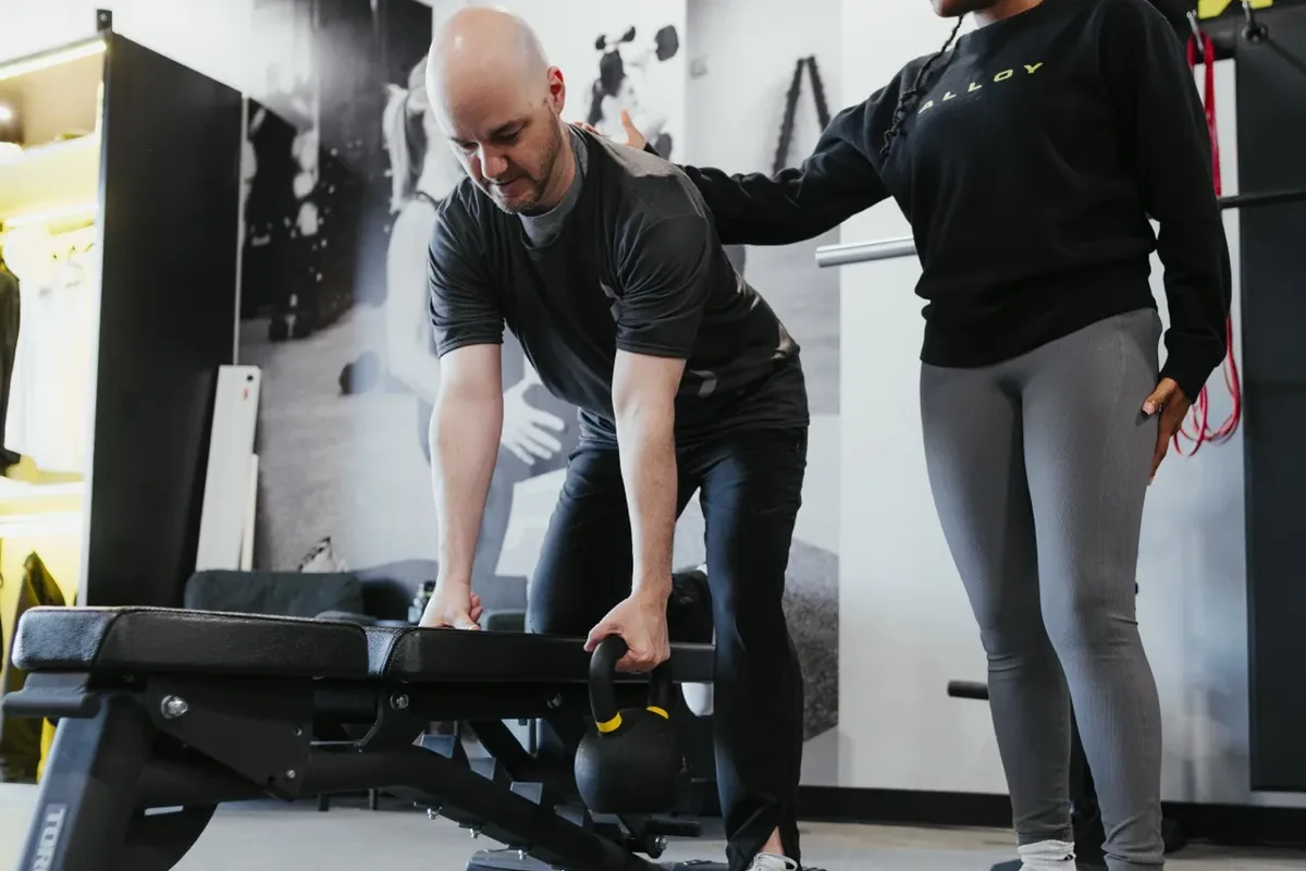 A coach guides a member setting up a kettlebell exercise at an adjustable bench during a session at Alloy Personal Training Middleton in Middleton, WI.