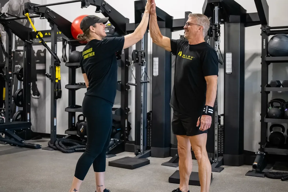 A coach and an older member share a high five in front of the cable machines at Alloy Personal Training Middleton in Middleton, WI.