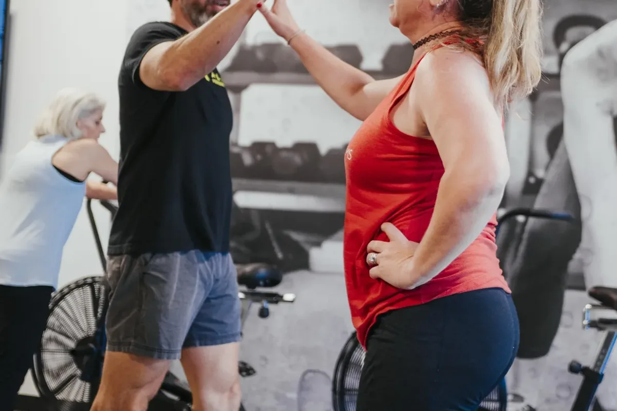 A coach high-fives a member after a set while another member rides a bike behind them at Alloy Personal Training Middleton in Middleton, WI.
